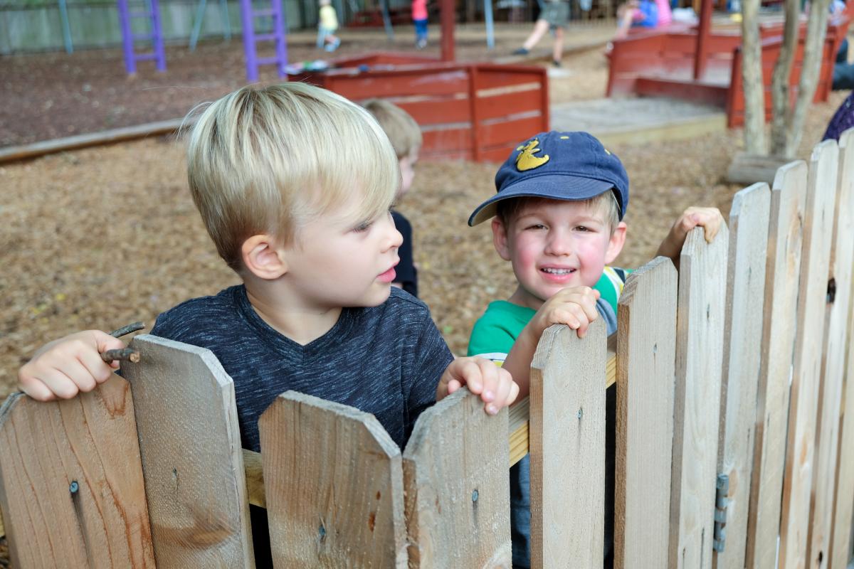 Two little boys look out from over a fence. One is wearing a blue hat and smiling at the camera.